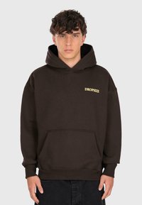 Jeune homme portant un sweat à capuche marron foncé avec une poche à l'avant et le texte jaune "DROPSIZE" sur la gauche de la poitrine, se tenant contre un fond uni.