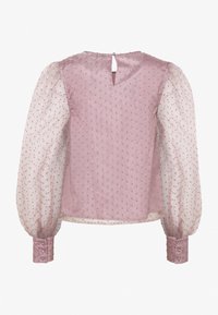 Blouse lilas avec manches longues transparentes à pois, poignets recouverts de tissu, et une petite fermeture à bouton en forme de trou à l'arrière de l'encolure.