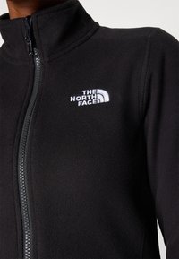 Zwarte fleecetak met een hoge kraag, voorzien van een voorsluiting met rits en een geborduurd wit logo op de linkerkant van de borst. Zachte textuur.