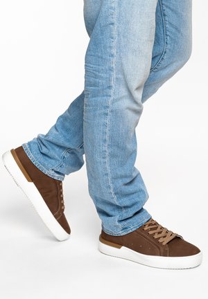 Persoon die lichtblauwe spijkerbroek en bruine sneakers met witte zolen draagt, staand tegen een effen witte achtergrond.