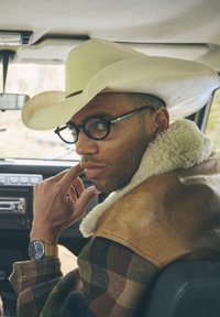 Καφέ δερμάτινο μπουφάν με γιακά από γούνα, καρώ φανελένιο πουκάμισο, λευκό καουμπόικο καπέλο, στρογγυλά γυαλιά και χρυσό ρολόι χειρός.