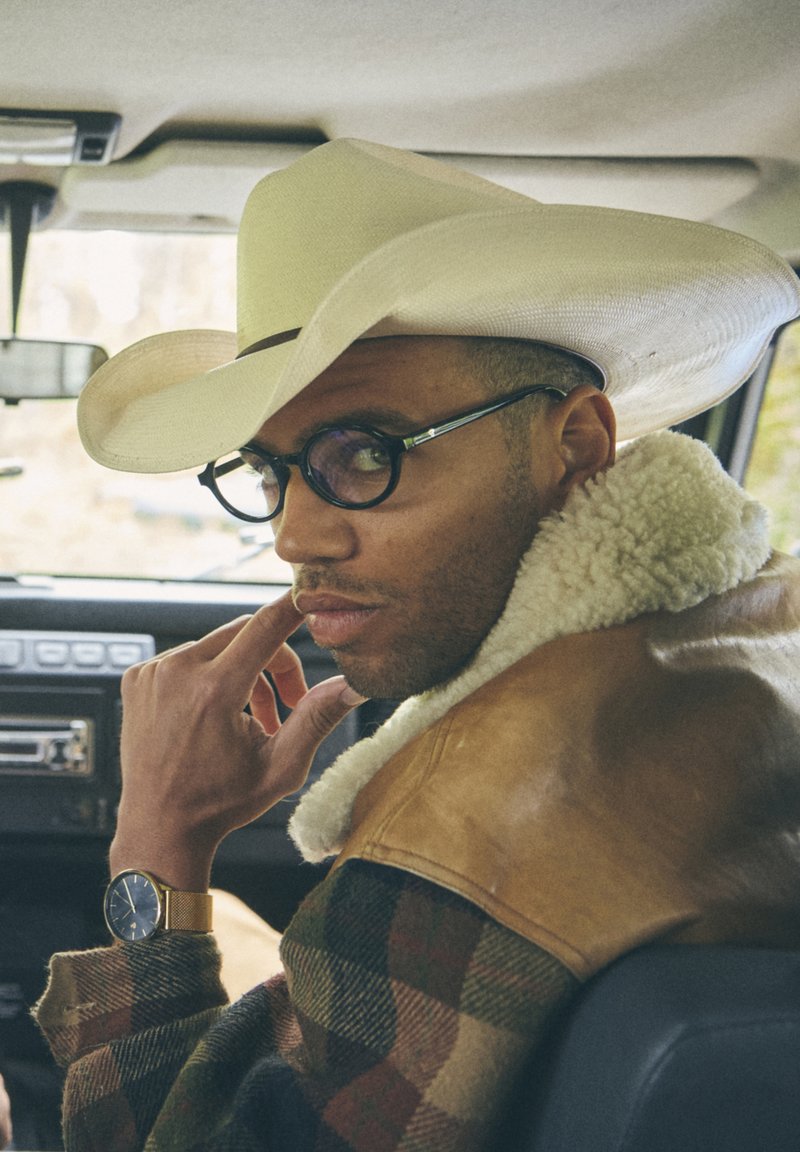 Καφέ δερμάτινο μπουφάν με γιακά από γούνα, καρώ φανελένιο πουκάμισο, λευκό καουμπόικο καπέλο, στρογγυλά γυαλιά και χρυσό ρολόι χειρός.