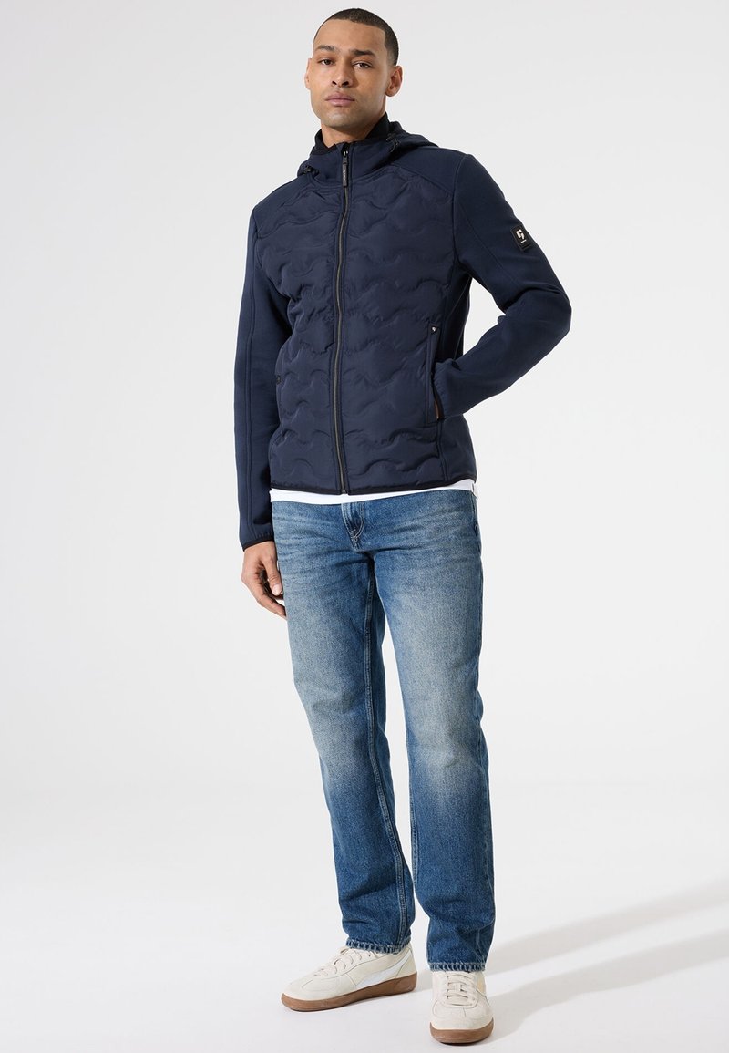 Veste à capuche bleu marine avec une texture matelassée, deux poches zippées et une coupe ajustée, associée à un jean bleu clair et des sneakers blanches.