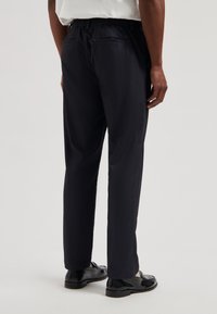 Mørkeblå bukser med elastisk midjebånd, to baklommer, glatt front, rette ben og en teksturert finish. Sorte sko fullfører antrekket.