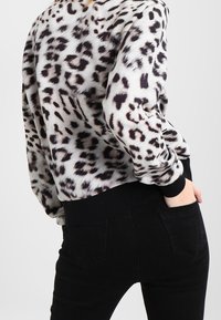 Persona vistiendo una camiseta de manga larga con estampado de leopardo en blanco y negro con puños negros, combinada con pantalones negros, mano en el bolsillo trasero.