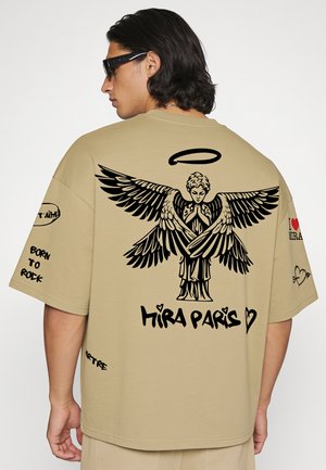 Mann trägt beige oversized T-Shirt mit schwarzer Engel-Grafik, ausgebreiteten Flügeln, Heiligenschein darüber und dem Text "Hira Paris" auf dem Rücken.