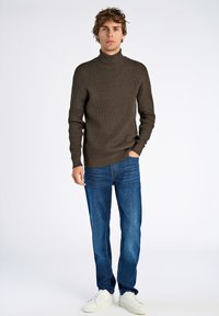 Braun strukturierter Rollkragenpullover, blaue Jeans und weiße Sneakers; Modell steht mit den Händen in den Taschen vor einem schlichten Hintergrund.