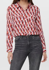 Camicia a maniche lunghe con stampa a zigzag rossa e bianca, con due tasche sul petto e una vestibilità comoda, abbinata a jeans grigi scuri a vita alta.