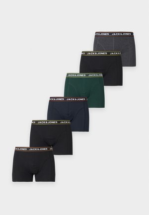 Homme mince à la peau foncée debout pieds nus, portant un boxer bleu marine Jack & Jones, regardant vers sa gauche sur un fond blanc uni.