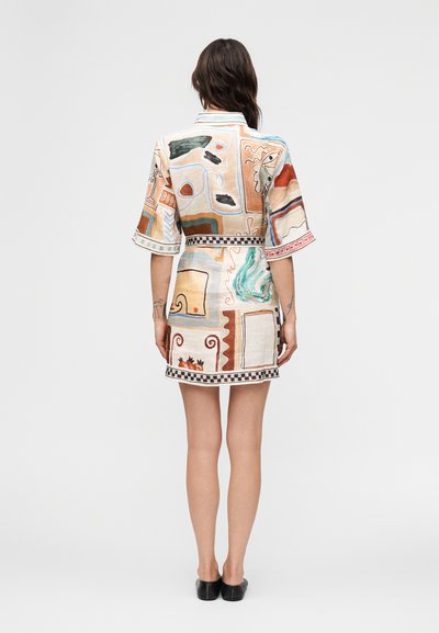 Γυναίκα με σκούρα μαλλιά φοράει κοντό φόρεμα με σχέδια και μανίκια ως τους αγκώνες, στέκεται μπροστά σε λευκό φόντο, με ορατά μαύρα παπούτσια χωρίς κορδόνια.