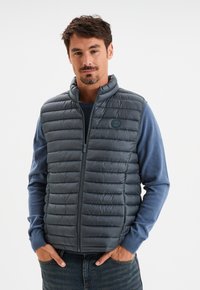 Grau gepolsterte Weste mit hochgeschlossenem Kragen und frontalem Reißverschluss. Besitzt eine gerippte Textur und ein Logobadge, getragen über einem blauen Langarmshirt.