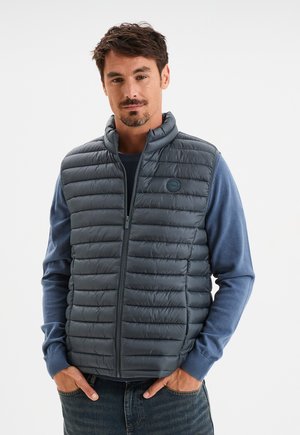 Gilet rembourré gris avec un col haut et une fermeture éclair à l'avant. Présente une texture côtelée et un badge logo, porté par-dessus une chemise à manches longues bleue.