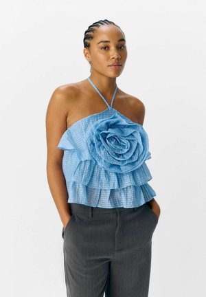 Haut halter bleu avec un grand détail de rose et des volants superposés. Fabriqué en tissu texturé. Associé à un pantalon gris.