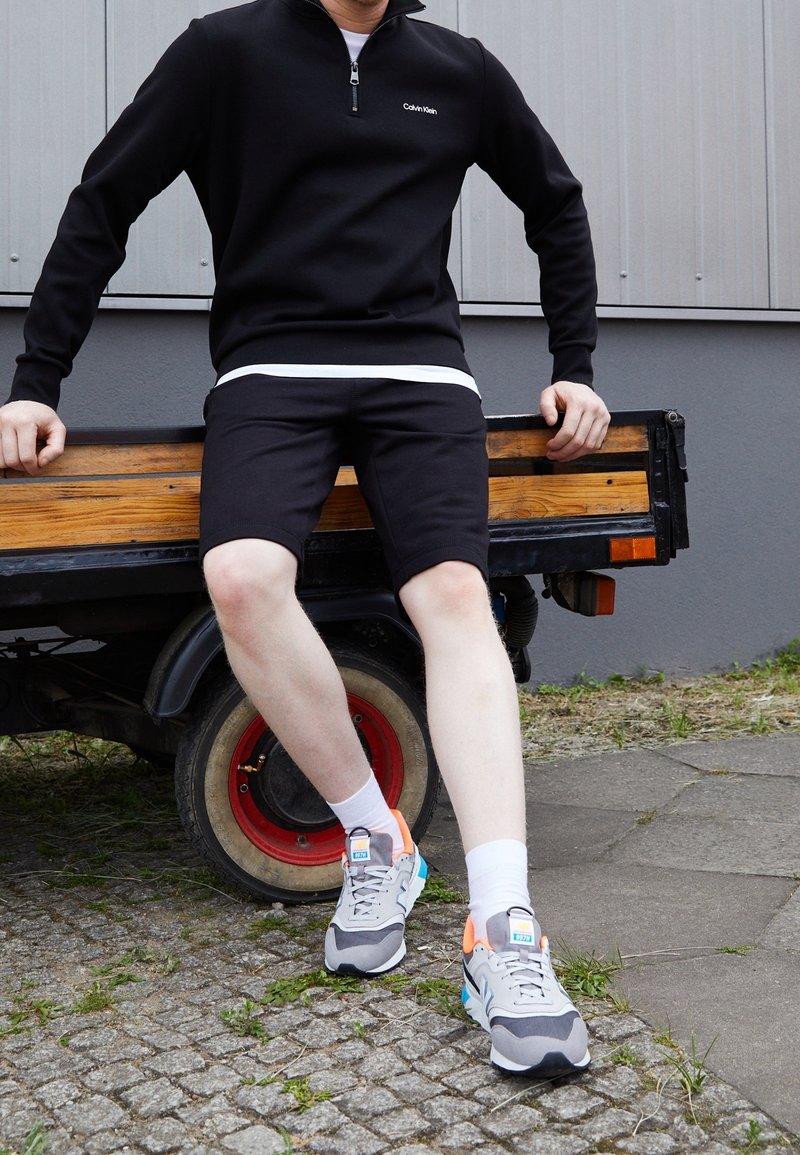 Personne portant une chemise noire à manches longues et un short est assise sur une remorque en bois, jambes tendues, portant des chaussettes blanches et des baskets grises avec des accents bleus et orange.