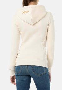 Cremefarbener Hoodie mit weicher Textur, ausgestattet mit einer Kapuze und gerippten Bündchen, akzentuiert durch ein goldfarbenes Logo auf der Rückseite. Kombiniert mit blauen Jeans.