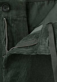 Zbliżenie na ciemnozielone spodnie sztruksowe z rozpiętym zamkiem błyskawicznym i widocznym czarnym guzikiem na pasie.