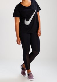 Musta Nike-paita, jossa on näyttävä hopeinen logo, yhdistettynä mustiin leggings-housuihin, joissa on verkko-ulkokäyttöä korostavia yksityiskohtia, sekä violetteihin urheilukenkiin.