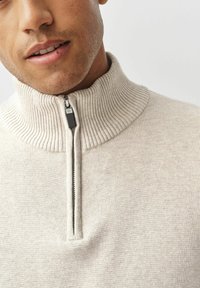 Bărbat îmbrăcat cu un pulover din tricot ribbed, bej, cu fermoar deschis parțial, având o bărbiță ușoară pe fața inferioară și buzele ușor deschise.