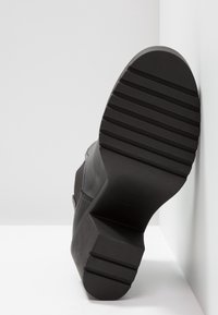 Botte noire avec talon épais et semelle rainurée, positionnée semelle contre un mur d'angle blanc et le sol.