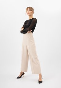 Top lungo nero trasparente a maniche lunghe abbinato a pantaloni beige a vita alta e gamba larga, con tacchi a punta neri caratterizzati da un'accenno dorato sul tacco.