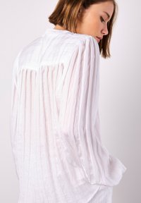 Weiße plissierte Bluse mit langen Ärmeln, strukturiertem Stoff und lockerer Passform. Besitzt eine dezente Rückenpasse und einen runden Halsausschnitt.