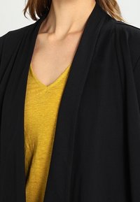 Svart kavaj med öppen frontdesign, bärs över en senapsgul V-ringad topp. Har mjukt tyg och avslappnad passform.