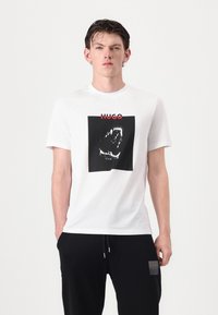 Hvit bomulls t-skjorte med et svart firkantet grafisk design med en stilisert munn og "HUGO"-tekst i rødt. Korte ermer og rund hals.