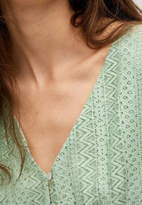 Blouse vert clair avec un motif en dentelle complexe, design en V et petits boutons sur le devant pour la fermeture.