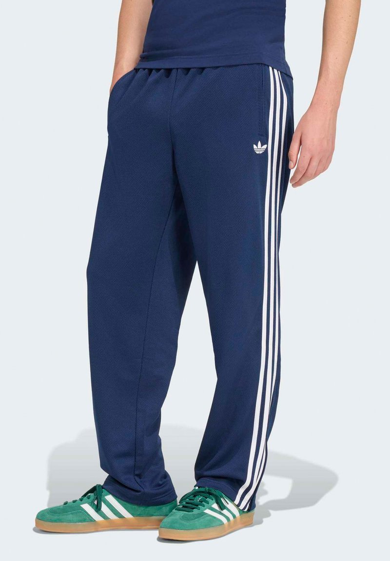 Osoba ubrana w granatowe spodnie dresowe Adidas z białymi paskami oraz zielone buty sportowe Adidas z białymi paskami, stojąca na tle jednolitego tła.