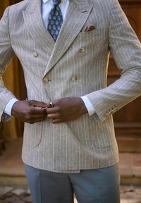 Blazer doppiopetto a righe marrone chiaro realizzato in tessuto texturizzato, con bottoni beige e fazzoletto da taschino, abbinato a pantaloni azzurri chiari.