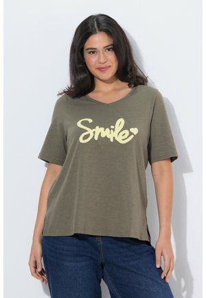 Frau mit langem dunklem Haar, die ein olivgrünes T-Shirt mit dem Schriftzug "Smile" und kleinem Herz trägt, posiert vor einem schlichten weißen Hintergrund.