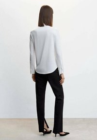 Weiße Bluse mit langen Ärmeln und abgerundetem Saum, kombiniert mit schwarzer, taillierter Hose mit seitlichen Schlitzen und schwarzen High Heels.