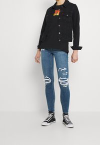 Chaqueta de mezclilla negra sobre una camiseta gráfica negra, combinada con pantalones vaqueros ajustados rasgados en azul y zapatillas negras de caña alta.