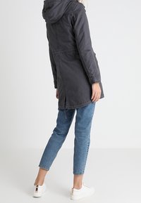 Černá kapucí parka se staženým pasem, dlouhými rukávy a bočními rozparky. Nositá přes modré džíny a bílé tenisky. Hladká textura, neformální design.