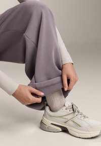 Světle fialové tepláky s ohrnutým lemem, kombinované se bílou a šedou teniskou. Materiál vypadá hladce a pružně.