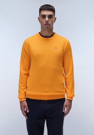 Maglione arancione in morbido tessuto lavorato a maglia con scollo rotondo e polsini a costine. Piccolo dettaglio del logo sul petto, indossato sopra pantaloni scuri.