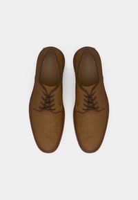 Chaussures à lacets en daim marron avec une finition lisse, des lacets fins et foncés, et une bordure cousue. Bout arrondi et design minimaliste.