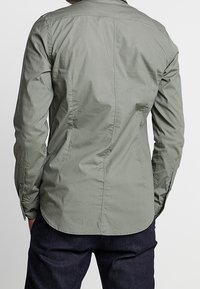 Jasnooliwkowa koszula z tkaniny o delikatnym splocie, z zapięciem na guziki przy kołnierzy, długimi rękawami i zaokrąglonym dołem, z subtelnymi detalami w szyciu.