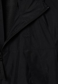 Giacca nera con zip, dal aspetto opaco, con fodera in rete e un discreto logo nero sul petto. Progettata per un'agevole stratificazione leggera.