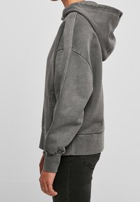 Osoba ubrana w wypłowiały szary sweatshirt z kapturem, z kieszenią typu kangurka i ściągaczami, w połączeniu z ciemnymi dżinsami, pokazana z boku.