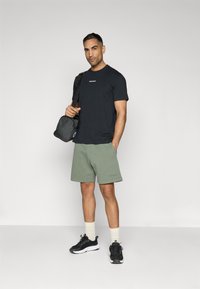 Black t-shirt with white logo, olive green shorts, black athletic sneakers, and cream-colored socks. Gray bag over right shoulder.