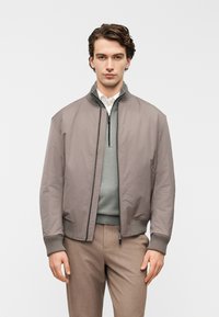 Γκρι μπουφάν τύπου bomber με ριμπ μανσέτες, κλείσιμο με φερμουάρ και λείο, ματ φινίρισμα. Φορεμένο πάνω από ένα ανοιχτό πράσινο πουλόβερ με φερμουάρ στο 1/4.