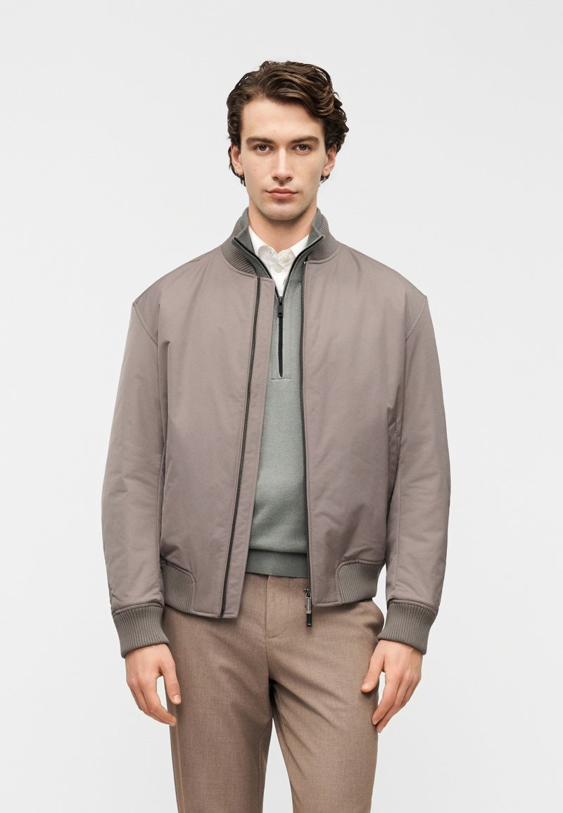 Γκρι μπουφάν τύπου bomber με ριμπ μανσέτες, κλείσιμο με φερμουάρ και λείο, ματ φινίρισμα. Φορεμένο πάνω από ένα ανοιχτό πράσινο πουλόβερ με φερμουάρ στο 1/4.