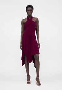 Γυναίκα με ασύμμετρο αμάνικο σκούρο κόκκινο φόρεμα χείλη και μπεζ ψηλοτάκουνα ανοιχτά παπούτσια, στέκεται μπροστά σε απλό λευκό φόντο.