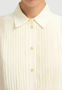 Crème-kleurige blouse met een klassieke kraag, voorzien van verticale plooien, stoffen knopen en een lichte, zachte textuur.