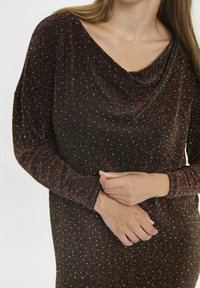 Moteris vilki ilgomis rankovėmis, rudos blizgančios suknelės su lankstymu kaklo linijoje, suspaudusi rankas ties juosmeniu.