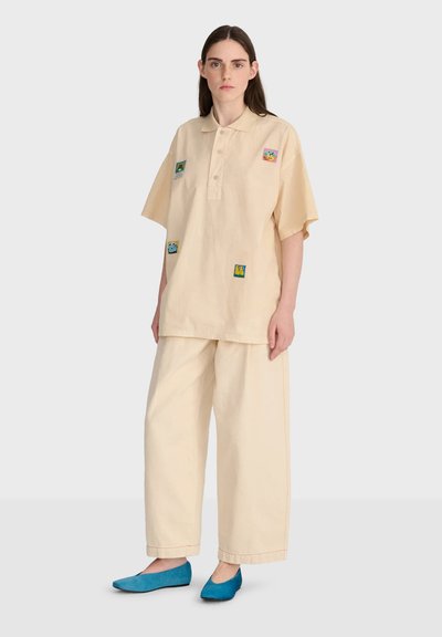 Femme portant un pantalon beige ample et une chemise beige à manches courtes avec des patches colorés, associée à des chaussures plates bleues, debout sur un fond uni.