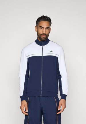 Marineblaues und weißes Zip-Up-Jackett mit Stehkragen; verfügt über ein horizontales Streifenmuster und ein kleines grünes Logo auf der Brust. Glatte Textur.