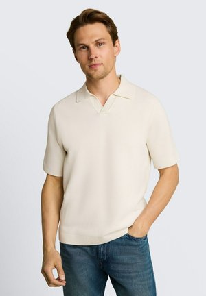 Jeune homme portant un polo en maille crème à manches courtes et un jean bleu, debout avec une main dans la poche devant un fond uni.