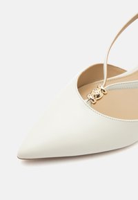 Chaussure plate blanche à bout pointu avec une fine bride à la cheville ornée d'une boucle décorative dorée sur fond clair.